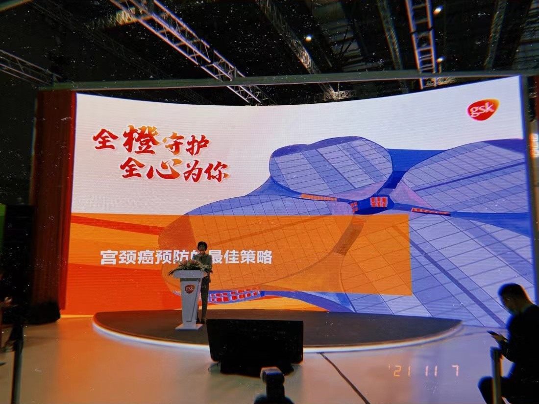 A speaker at a podium in front of a very wide curved LED wall reading, in Chinese, the best strategy for cervical cancer prevention.