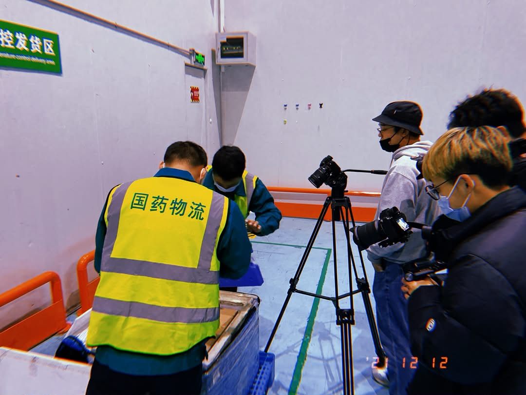 A temperature-controlled pharmaceutical dispatch bay: warehouse staff in high-visibility vests packing boxes while three photographers film them.
