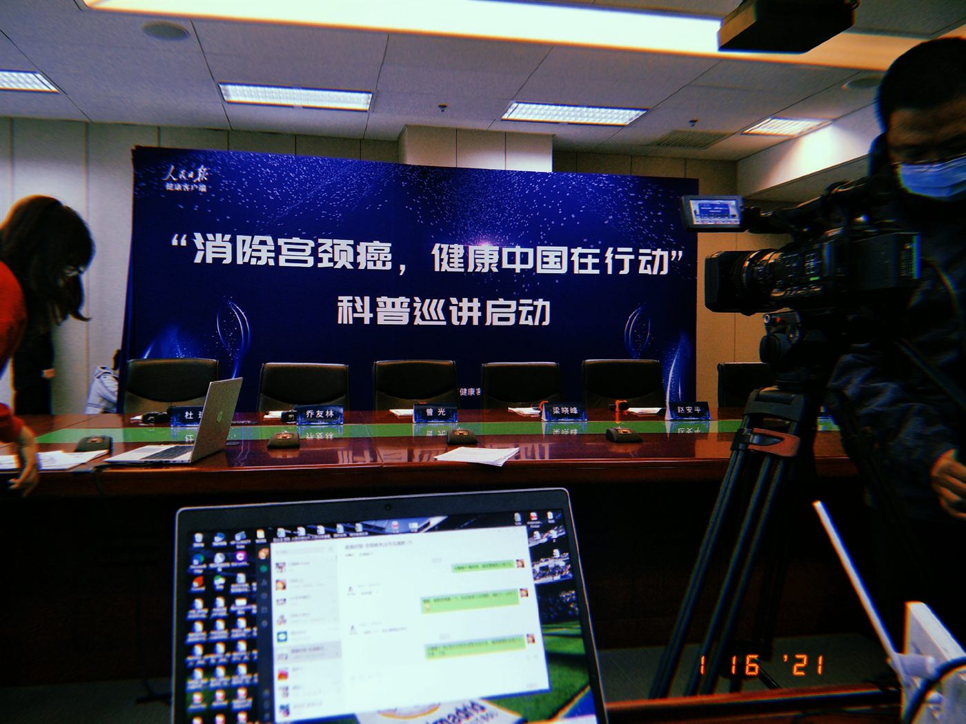 An empty press conference table at People's Daily, five name cards set out in front of a campaign backdrop, with a broadcast camera on the right.