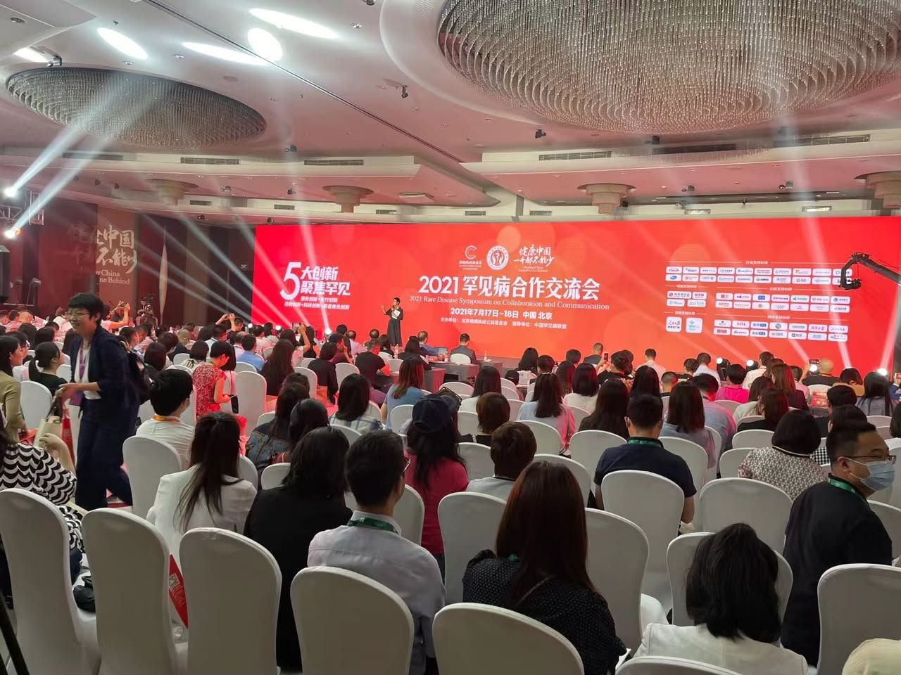 A full conference hall under stage lighting, facing a wide red backdrop listing dozens of partner organisation logos.