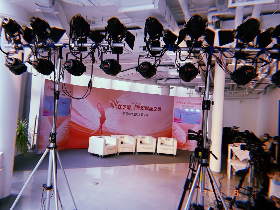 An empty television studio: a rig of about a dozen film lights overhead, four white armchairs facing a pink three-panel backdrop.